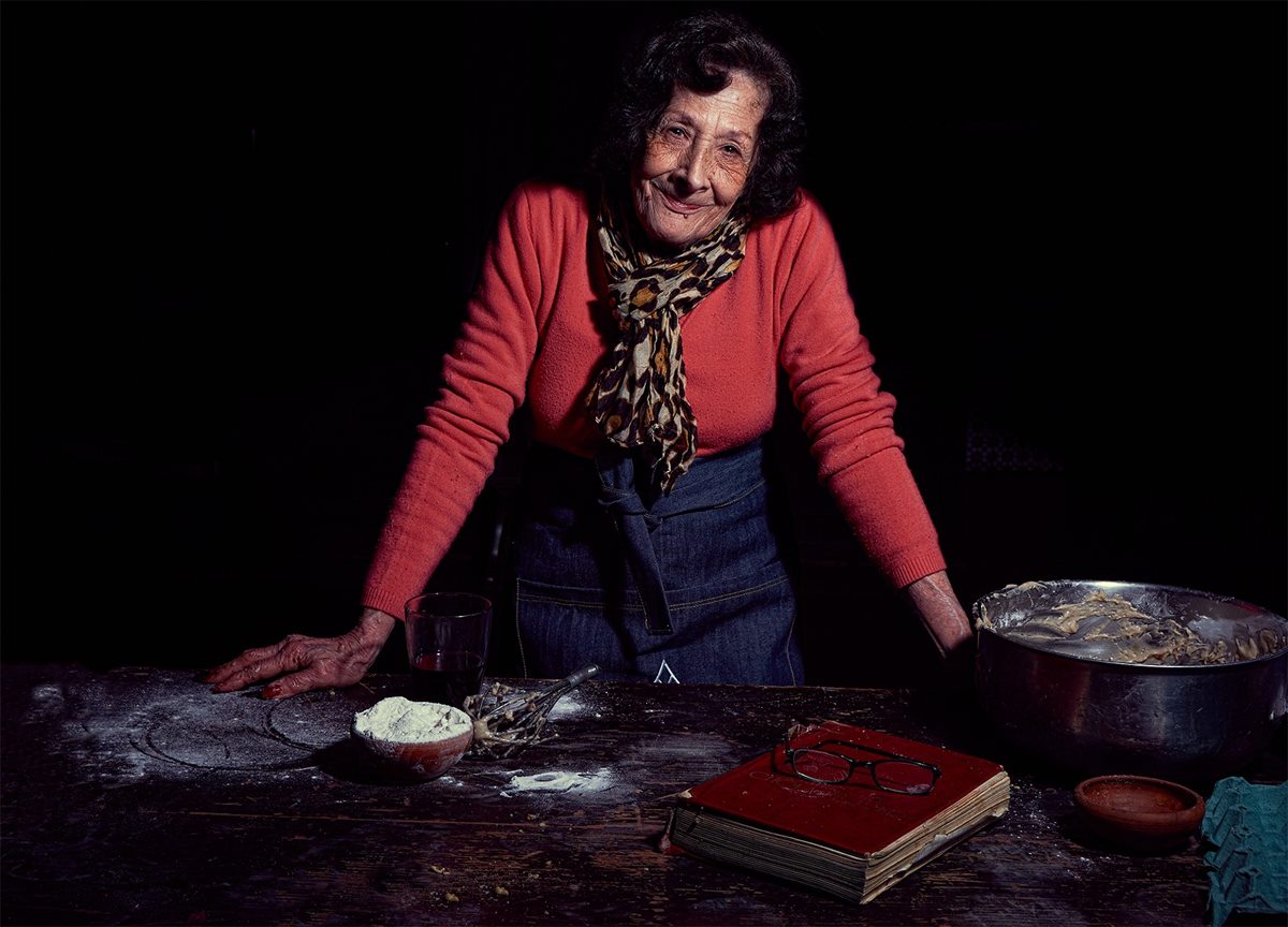 Blanca Lanzy a color, sonriendo a cámara, apoyada en una mesa de madera, con pulóver rojo y delantal de jean