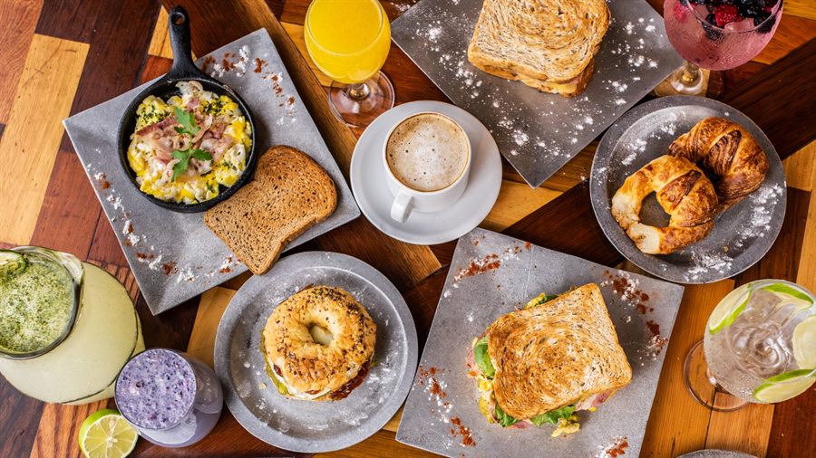 Mesa de desayuno con café con leche, medialunas, tostados, revuelto con panceta, jugo de naranja, limonada y licuado